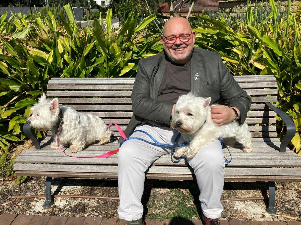 Michael and his two west highland terrier therapy dogs sit on a park bench together